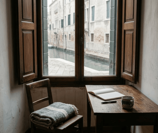 Wooden desk below a tall window overlooking a Venetian canal in late autumn light, with an open notebook, a ceramic mug and a folded wool throw on a chair