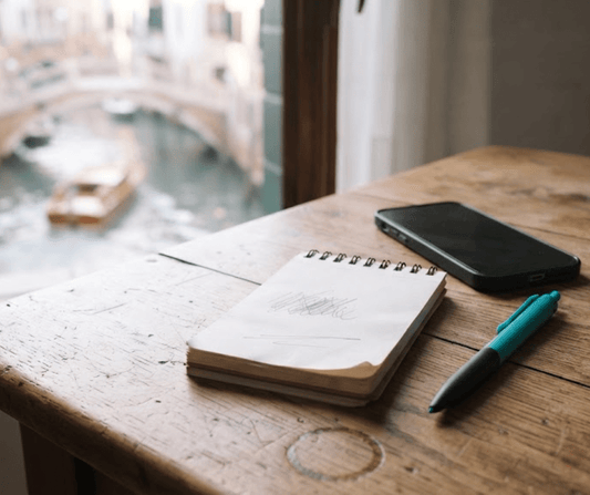 Notepad, smartphone and teal pen on a wooden desk by a window over a Venetian canal, for Airbnb host fee repricing