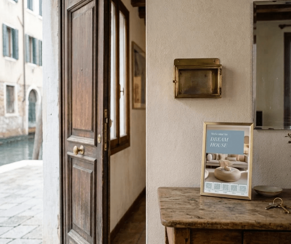 Open wooden door onto a Venetian canal, with a framed welcome sign showing five QR codes on a wooden console beside a ceramic dish and keys, and a brass wall box above.